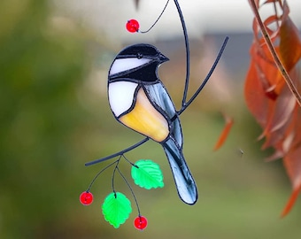 Puede incluir: Un pájaro de vidrio coloreado con detalles negros, blancos, amarillos y grises posado en una rama con hojas verdes y bayas rojas.