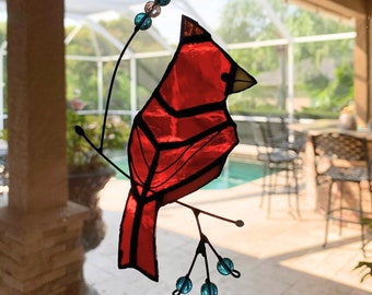 May include: A stained glass cardinal bird ornament with red and black accents, perched on a branch with blue glass beads. The ornament is hanging from a window.