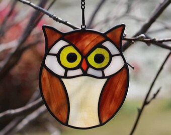 May include: A stained glass owl ornament with brown, white, and yellow accents. The owl has large, round eyes and a detailed design.