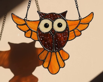 May include: A stained glass owl with brown and orange glass, hanging from a black chain. The owl has large black eyes and is in flight.