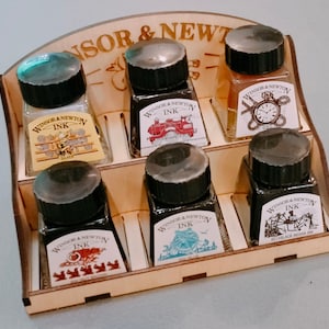 Könnte beinhalten: Ein Holzdisplay mit sechs Flaschen Winsor & Newton Tinte. Die Flaschen sind in zwei Reihen mit je drei Flaschen angeordnet. Jede Flasche hat einen schwarzen Verschluss und ein Etikett mit dem Winsor & Newton Logo und dem Wort "INK".