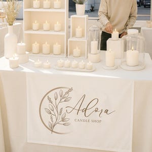 Puede incluir: Un puesto con velas encendidas de varios tamaños, incluyendo velas de pilar y velas de té, sobre una mesa blanca. La mesa tiene una pancarta con el texto "Adora Candle Shop" y un diseño floral. Una persona está encendiendo una vela.
