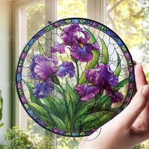 May include: A round stained glass panel featuring purple iris flowers with green leaves. The panel has a black metal frame and is held in front of a window. The iris flowers are in full bloom, with detailed petals and vibrant colors.