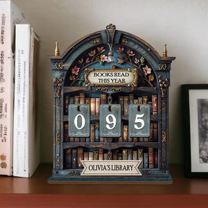 May include: A decorative wooden bookshelf-shaped calendar with the text "BOOKS READ THIS YEAR" and "OLIVIA'S LIBRARY". The calendar displays the number "095" with individual number tiles. The design features floral accents and a blue and brown color scheme.