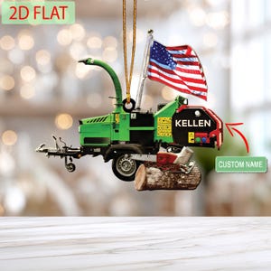 May include: A green and red wood chipper ornament with an American flag and the name "KELLEN" on the side. The ornament is hanging by a gold string and has the text "2D FLAT" on the top left.