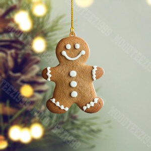 K&ouml;nnte beinhalten: Ein Lebkuchenmann-Weihnachtsschmuck mit wei&szlig;en Zuckergussdetails und einem l&auml;chelnden Gesicht. Der Schmuck h&auml;ngt an einem goldenen Faden vor einem verschwommenen Hintergrund aus Weihnachtslichtern und Tannennadeln.