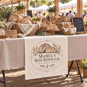 May include: A beige linen table runner featuring a circular graphic with two loaves of bread, wheat stalks, and the text Mabel's Sourdough, Natural Fermented Bread, Small Batch, Baked Fresh, displayed on a table with baskets of sourdough bread.