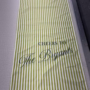 May include: A rectangular green and white striped table runner. The text "CHEERS TO The Bryants" is printed in black script. The runner is made of fabric and is laying flat on a surface.