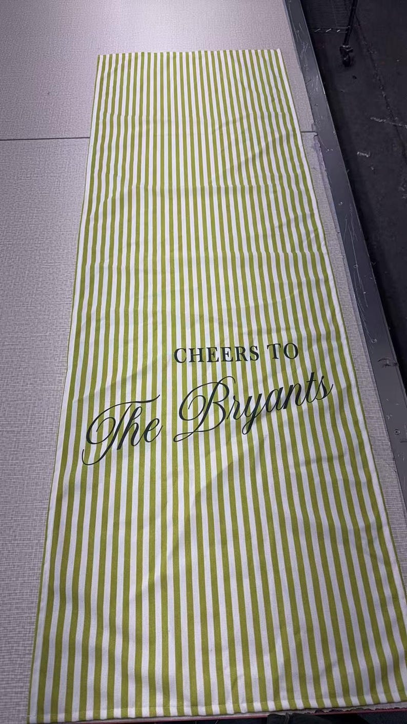 May include: A rectangular green and white striped table runner. The text "CHEERS TO The Bryants" is printed in black script. The runner is made of fabric and is laying flat on a surface.
