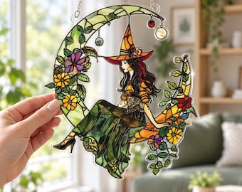 May include: A stained glass suncatcher shaped like a crescent moon featuring a witch with long dark hair wearing an orange hat and dress. The moon is decorated with green leaves and colorful flowers in shades of yellow, purple, and red.