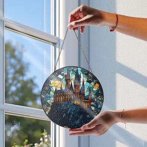 Könnte beinhalten: Runde Glasfensterkunst mit einer Burgszene unter einem Nachthimmel. Das Kunstwerk wird von einer Kette gehalten und hat einen schwarzen Rand. Die Burg ist braun und goldfarben, vor einem blauen, gelben und türkisfarbenen Hintergrund.