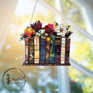 May include: A stained glass window hanging with a colorful depiction of a stack of books with flowers on top. The books are made of stained glass with different colors and patterns. The flowers are red, yellow, and white.