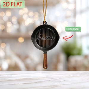 May include: A black cast iron skillet ornament with a wooden handle and vertical grill lines. The word "Name" is written in cursive across the skillet. A gold string is attached for hanging. The text "2D FLAT" is in a green rectangle.