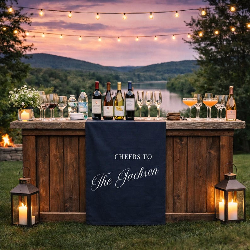May include: Outdoor bar setup with wine bottles, glasses, and cocktails. A navy blue cloth with the text "Cheers to The Jackson" hangs on the front of the wooden bar. Lanterns with lit candles flank the bar, with string lights overhead.