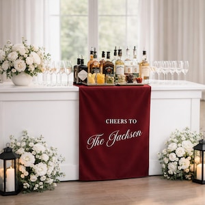 May include: A white bar setup with various liquor bottles, glasses, and appetizers. A burgundy runner with the text "Cheers to The Jackson" is draped over the bar. White floral arrangements and lanterns with candles flank the bar.