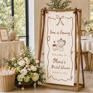 May include: A vertical fabric banner with brown stripes and a brown border, displayed on a wooden A-frame stand with brown ribbons. The banner features the text "love is brewing", a coffee pot and cup illustration, "WELCOME TO Olivia's Bridal Shower", and "MARCH 14, 2026".