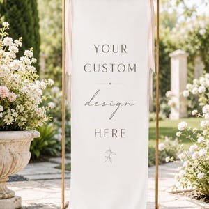 May include: A tall, rectangular white fabric banner suspended from a thin gold metal frame. The banner features the text "YOUR CUSTOM design HERE" in a mix of serif and script fonts, with a small leaf illustration at the bottom and pink ribbons.