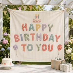 May include: A cream-colored fabric banner with thin vertical stripes featuring the text HAPPY BIRTHDAY TO YOU in multi-colored, patterned fabric letters. The banner includes a cake graphic with three candles and two balloon illustrations.
