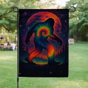 Puede incluir: Una bandera de jardín con una silueta de Bigfoot teñida con tinte frente a una luna naranja y roja. El diseño incluye un cielo estrellado y una silueta de bosque. La bandera se muestra al aire libre.