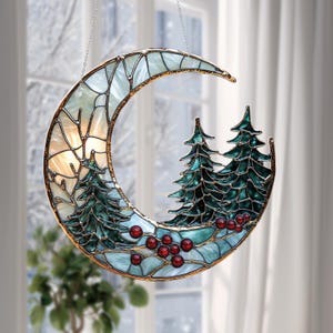 May include: Stained glass crescent moon with a winter forest scene. The moon features a light blue and white mosaic design with a gold-colored border. Two green pine trees and red berries are inside the moon's curve, creating a winter holiday theme.
