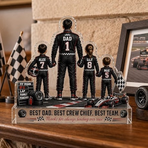 May include: A clear acrylic racing-themed family figurine. The figurine features a father figure and three children in racing suits, with a race car and checkered flags. The base reads "Best Dad. Best Crew Chief. Best Team."