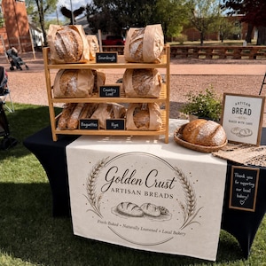May include: A display of artisan breads, including sourdough, country loaf, baguettes, and rye, presented on a wooden tiered shelf. The table features a sign that reads "Golden Crust Artisan Breads" and "Artisan Bread Baked With Care."