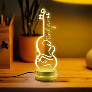 Könnte beinhalten: Ein gelbes LED-Nachtlicht in Form einer Geige mit dem Wort "Symphonie" darauf. Das Licht steht auf einem Holzsockel.