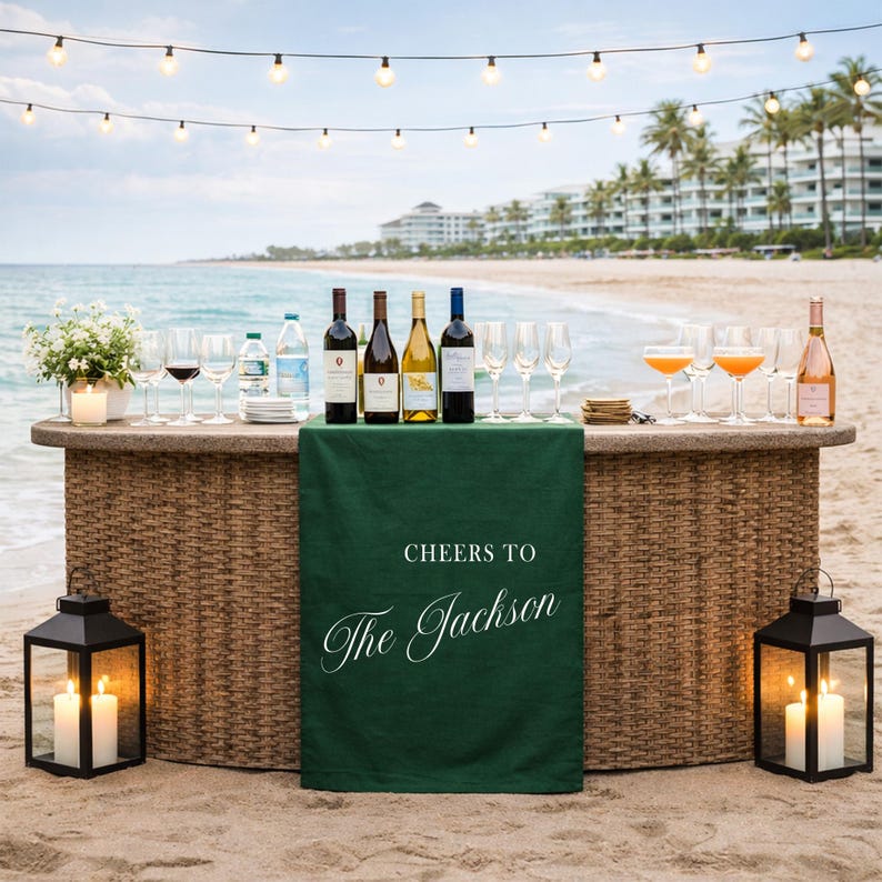 May include: A beachside bar setup with wine bottles, glasses, and cocktails. A green cloth with the text "Cheers to The Jackson" drapes the front. Two black lanterns with lit candles flank the bar. String lights hang overhead.
