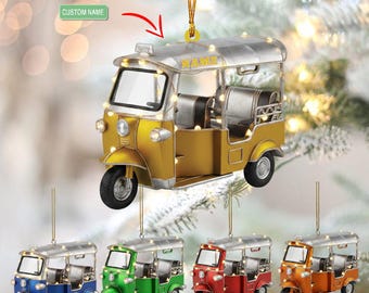 May include: A yellow and silver tuk-tuk ornament with the word "NAME" on top. The ornament has a string for hanging and is surrounded by fairy lights. Additional ornaments in blue, green, red, and orange are also shown.