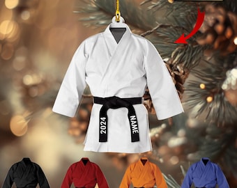 May include: A white martial arts uniform ornament featuring a black belt tied around the waist. The belt is printed with the text 2024 and NAME. A gold string is attached to the top for hanging. Additional uniform colors include black, red, orange, and blue.