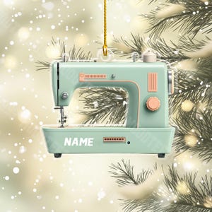 May include: A light green sewing machine ornament with a white "NAME" inscription. The ornament is hanging from a gold string and is surrounded by a blurred background of a Christmas tree and lights.