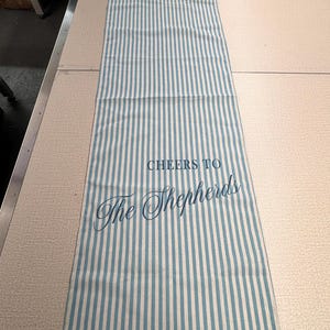 May include: A rectangular, striped kitchen towel with vertical blue and white stripes. The text "CHEERS TO The Shepherds" is printed in blue script across the center of the towel. The towel is lying flat on a light-colored surface.