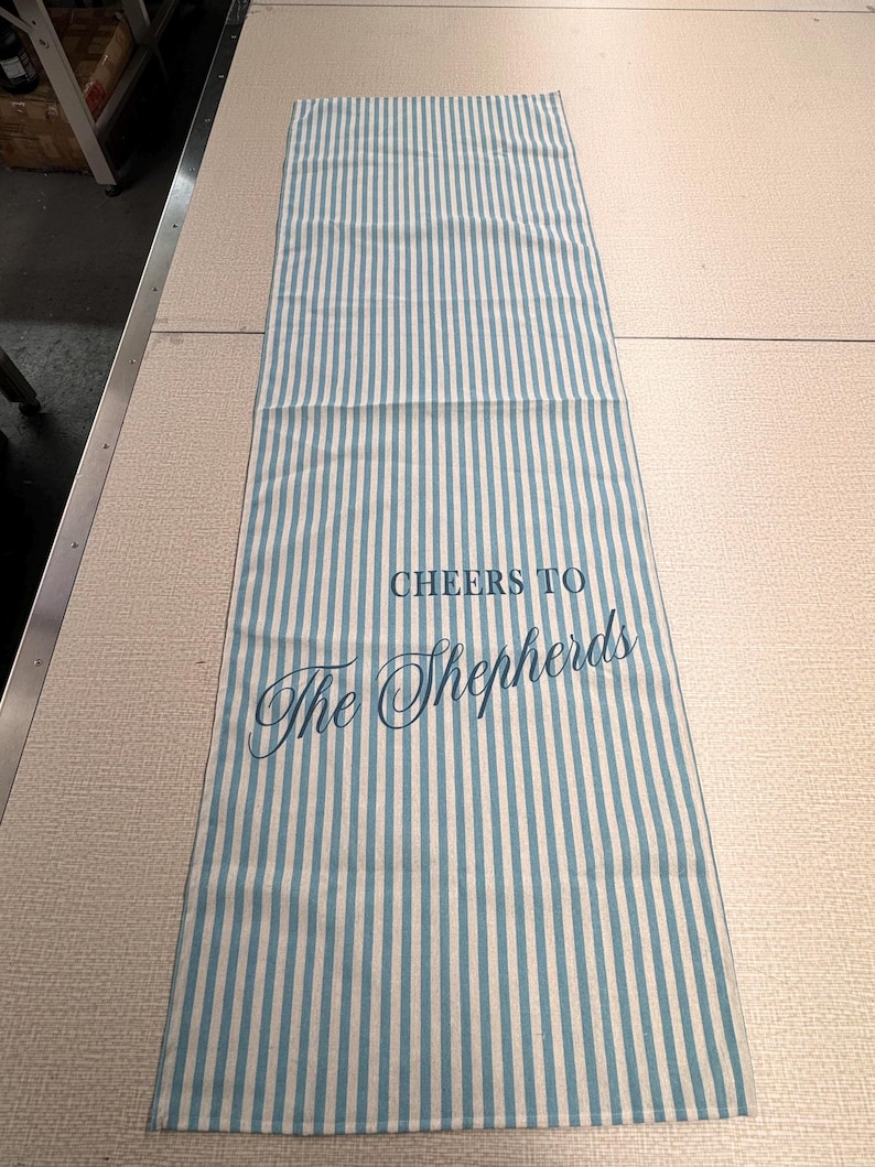 May include: A rectangular, striped kitchen towel with vertical blue and white stripes. The text "CHEERS TO The Shepherds" is printed in blue script across the center of the towel. The towel is lying flat on a light-colored surface.
