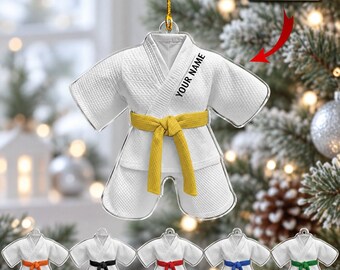 May include: A white martial arts uniform ornament featuring a yellow belt and the text YOUR NAME printed on the chest. The ornament hangs from a gold string and is shaped like a gi. Additional ornaments with orange, black, red, blue, and green belts are shown below.