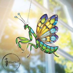 Puede incluir: Un adorno colgante de vidrio coloreado que representa una mantis religiosa con alas de mariposa. La mantis es verde con detalles dorados, y las alas son una mezcla de amarillo, azul y morado.