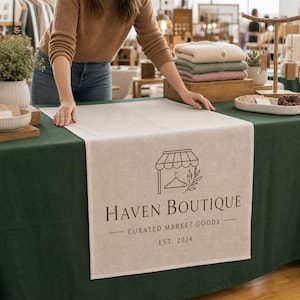 May include: A rectangular beige linen table runner featuring a dark brown printed logo of a boutique storefront with a hanger and leafy branch. The text reads Haven Boutique, Curated Market Goods, and Est. 2024, centered on the fabric surface.