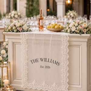 May include: A cream-colored fabric table runner featuring intricate white lace trim along the edges. The center of the runner is printed with the black text THE WILLIAMS and Est. 2026. The runner is draped over a light-colored wooden table.