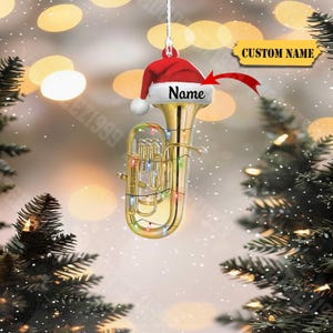 May include: A gold-colored tuba Christmas ornament with a Santa hat and string lights. The hat has the word "Name" on it. A banner with the words "CUSTOM NAME" is also present. The ornament hangs from a white string.