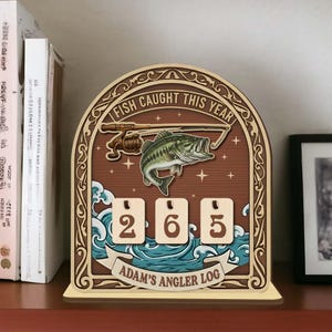 May include: Wooden fishing-themed counter with the text "FISH CAUGHT THIS YEAR" and "ADAM'S ANGLER LOG". The counter displays the number 265. Features a fish, fishing rod, and water wave design.
