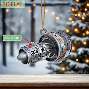 May include: A silver jet engine Christmas ornament with the text "TCCS, INC." and a space for a custom name. The 2D flat ornament hangs from a gold string. The background shows a snowy scene and Christmas lights.
