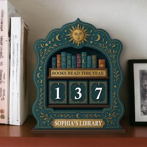 May include: A teal wooden perpetual calendar with a sun and moon design, featuring the text "BOOKS READ THIS YEAR" and the numbers "137". The calendar has a shelf with miniature book illustrations and the words "SOPHIA'S LIBRARY" at the bottom.
