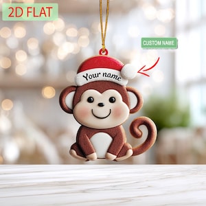 May include: A brown monkey Christmas ornament wearing a red Santa hat with a white pom-pom. The hat has the text "Your name" on it. The ornament is hanging from a gold string. The words "2D FLAT" are in the top left corner.