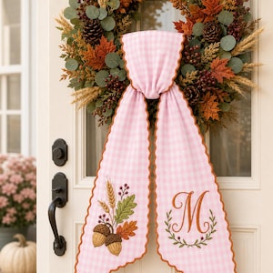 Puede incluir: Un colgador de puerta de tela de cuadros vichy rosa y blanco con bordes festoneados y ribete color &oacute;xido. Un lado presenta trigo, hojas de roble y bellotas bordadas, mientras que el otro muestra una letra M bordada dentro de una corona.