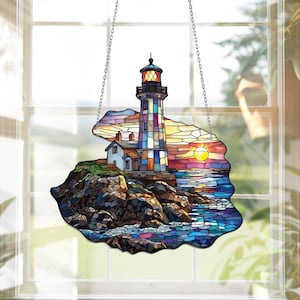 Puede incluir: Una ventana de vidrio emplomado colgada con una imagen colorida de un faro en una costa rocosa con una puesta de sol en el fondo.