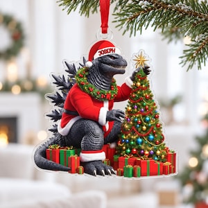 May include: A clear acrylic Christmas ornament featuring a black Godzilla character wearing a red Santa suit and hat with the name JOSEPH printed on the brim. The figure is decorated with a green wreath and is placing a gold star on a Christmas tree.