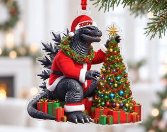 May include: A clear acrylic Christmas ornament featuring a black Godzilla character wearing a red Santa suit and hat with the name JOSEPH printed on the brim. The figure is decorated with a green wreath and is placing a gold star on a Christmas tree.