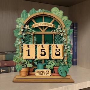 May include: Wooden bookshelf decor with a window design, featuring the text "BOOKS READ THIS YEAR" and the numbers "158". The decor includes potted plants, a watering can, and the text "SOPHIA'S LIBRARY".