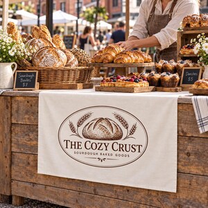 May include: A bakery stall display featuring a variety of sourdough boules in a wicker basket, croissants, and berry tarts. A cream-colored fabric banner hangs in front with the brown oval logo THE COZY CRUST SOURDOUGH BAKED GOODS.