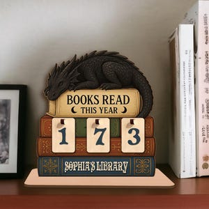 May include: A wooden book-themed decor piece with a black dragon resting on a stack of books. The books display the text "BOOKS READ THIS YEAR" and "SOPHIA'S LIBRARY". The numbers "173" are displayed on small cards.