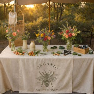 May include: A table display featuring bouquets of colorful flowers in glass vases and ceramic containers. The table is covered with a beige tablecloth with the words "ORGANIC SWEET HONEY" and a bee illustration. Scissors, twine, and other supplies are arranged on the table.